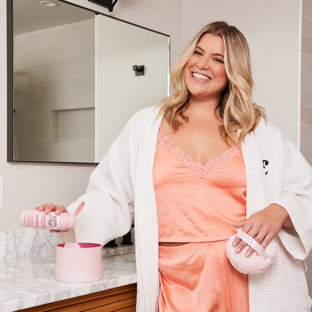 Smiling woman in a robe at a marble bathroom vanity holding Happy Curves Comfort Powder and the Comfort Puff, with the open puff tin on the counter